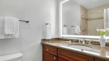 Un baño con inodoro blanco, lavamanos con encimera de granito, un espejo grande y armarios de madera.