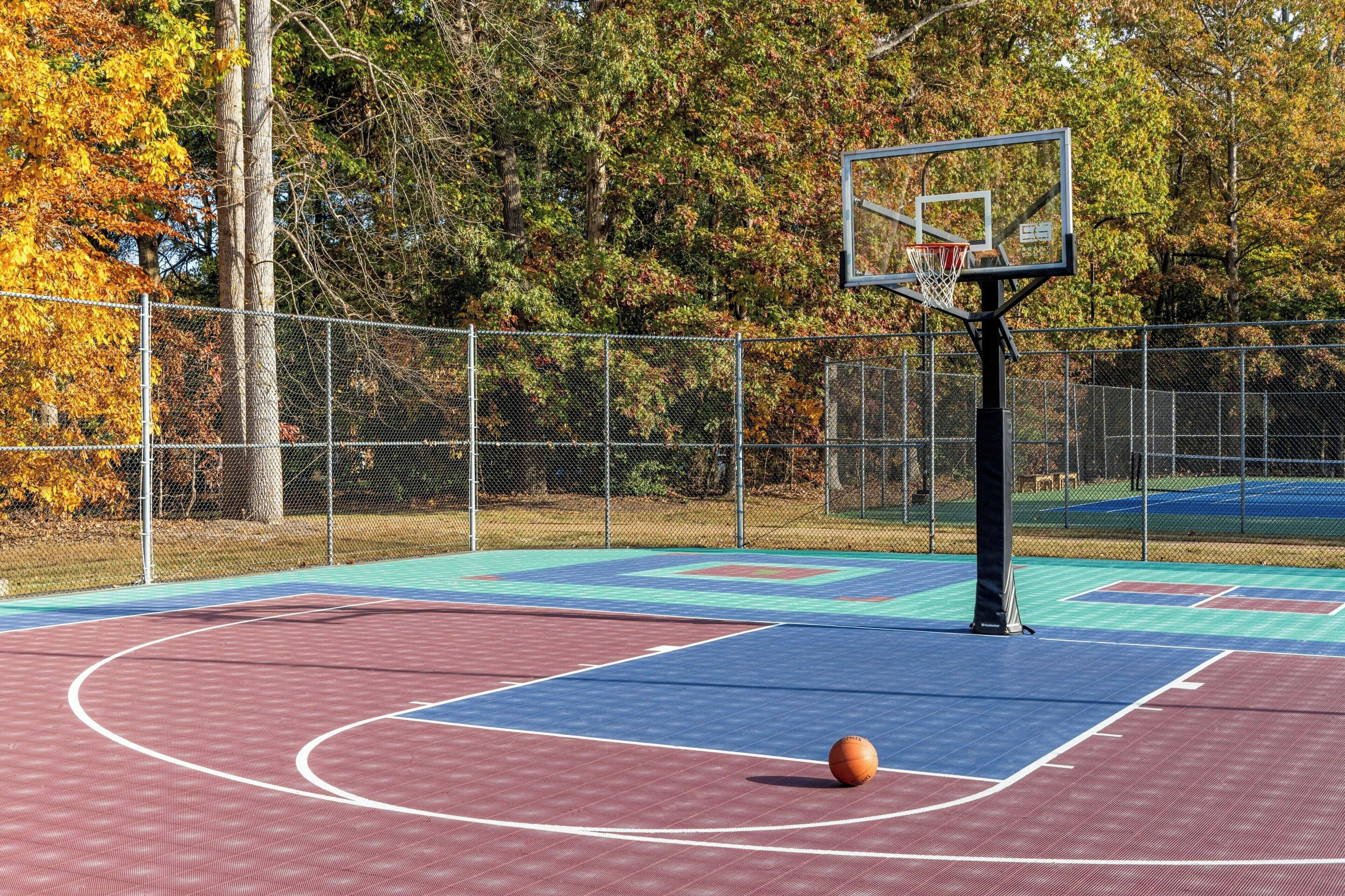 An outdoor basketball court with a chain-link fence, trees in the background, and a basketball hoop.