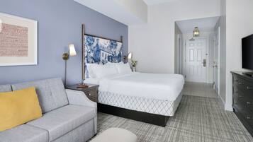 A hotel room with a bed, a sofa, a nightstand, a lamp, a TV, and a framed picture on the wall.