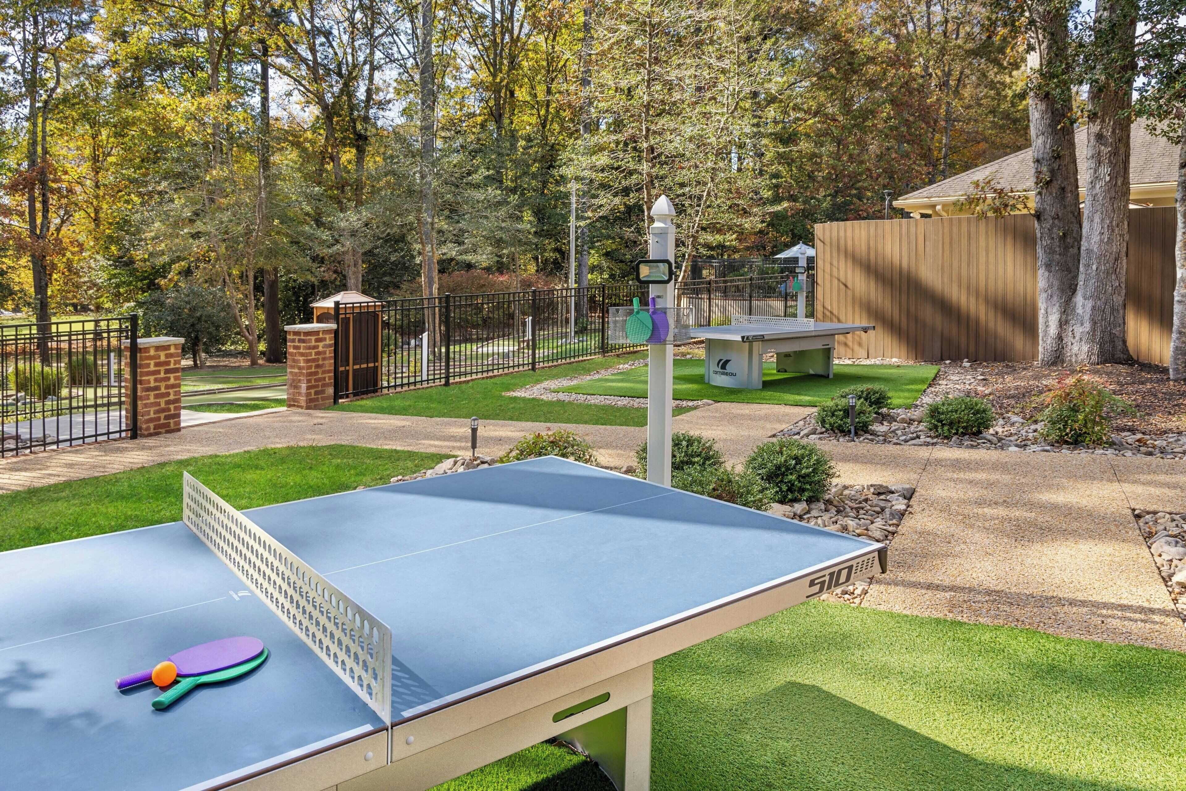 Mesa de ping pong al aire libre con una cerca, árboles y un banco en un entorno similar a un parque.