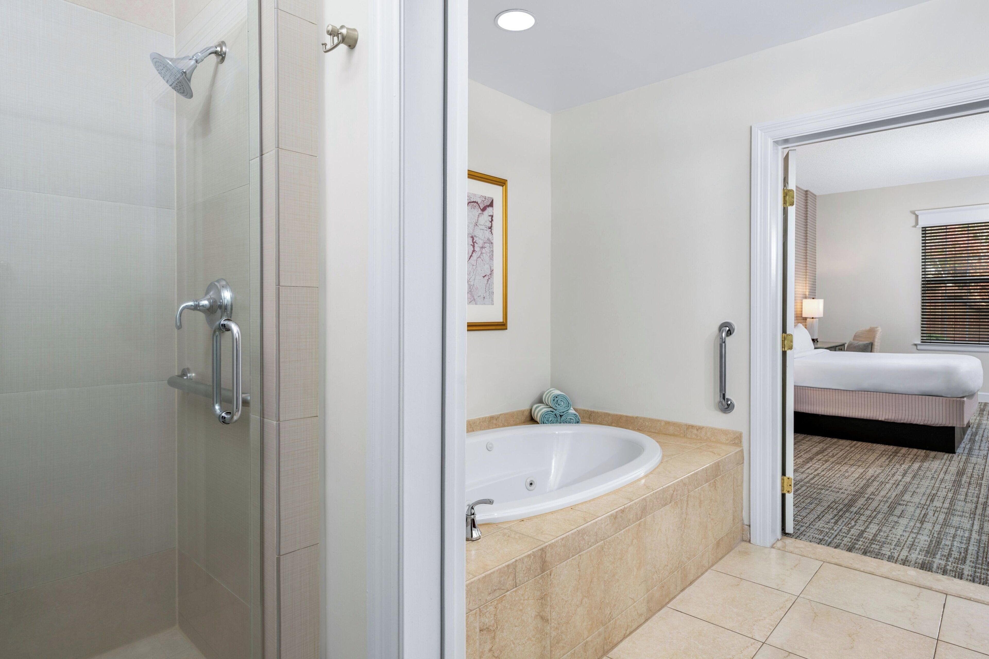 Un baño moderno con cabina de ducha de vidrio, una bañera independiente y una cama visible en la habitación contigua.
