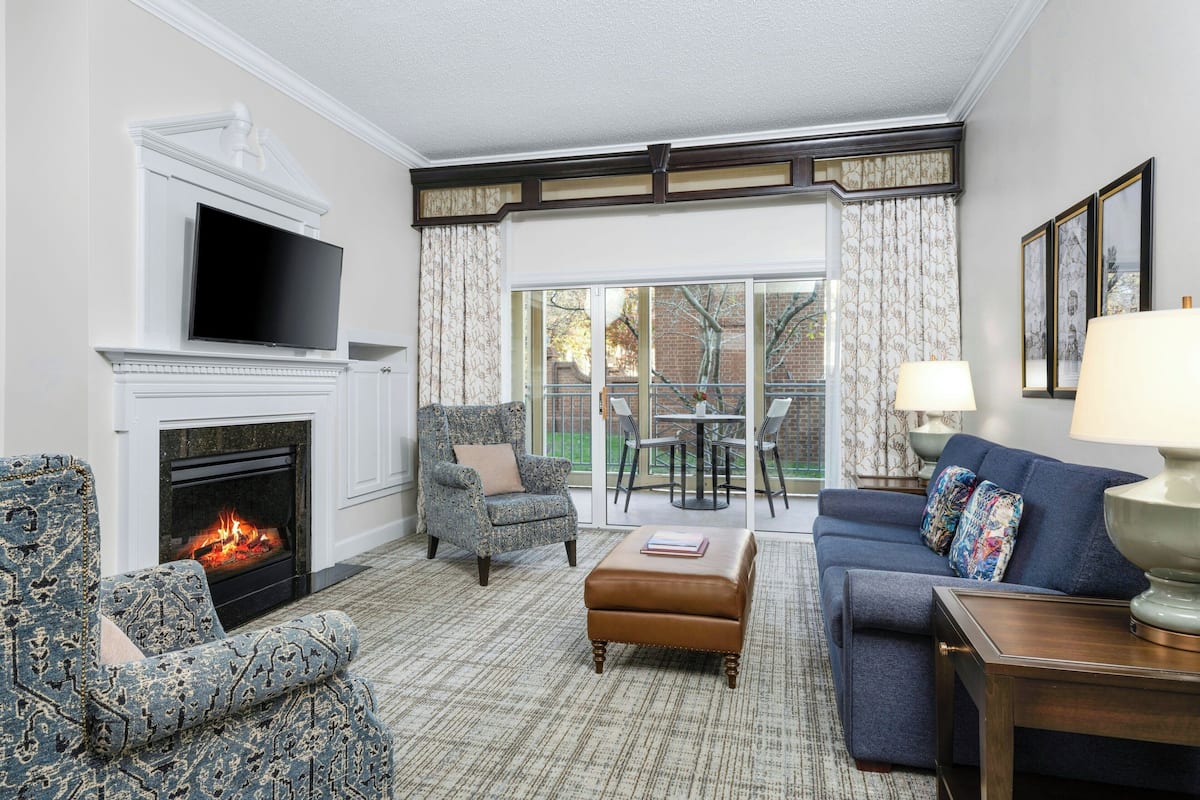 A living room with a fireplace, a television, a sofa, an ottoman, and a table with a lamp.