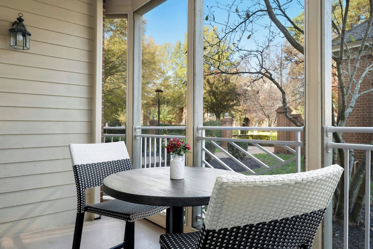 A covered patio with a round table and two chairs, a vase with flowers, and a view of trees and a brick building.