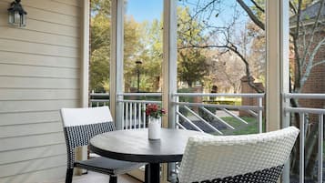 A covered patio with a round table and two chairs, a vase with flowers, and a view of trees and a brick building.