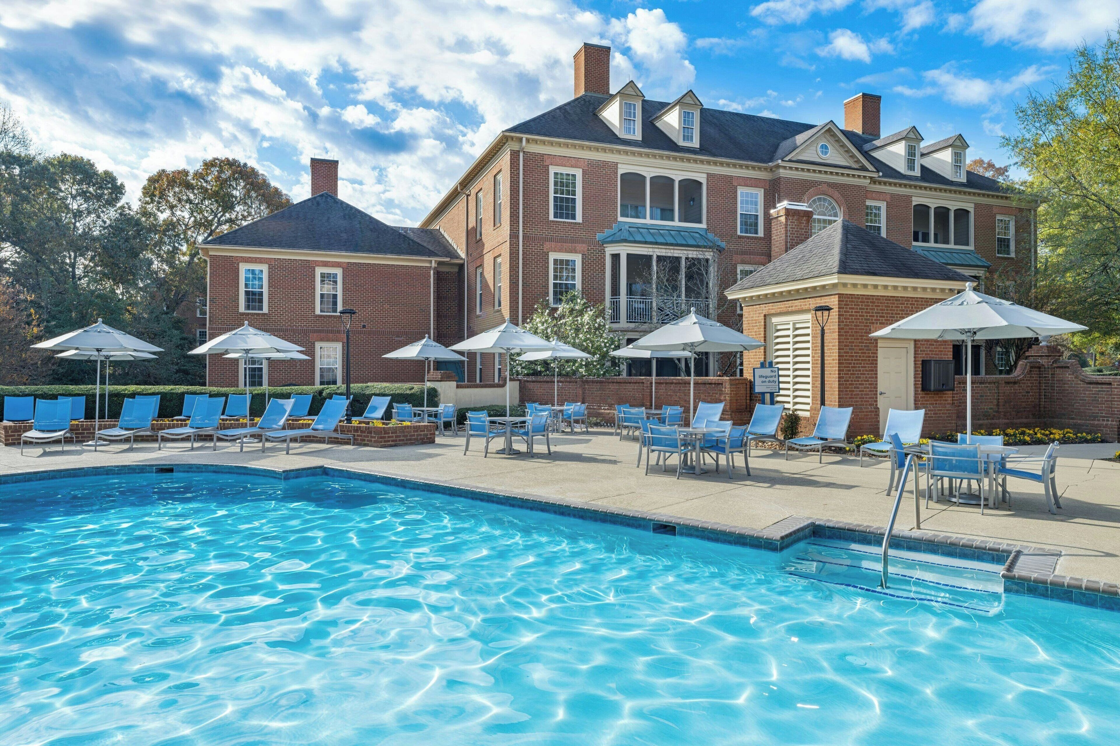 A swimming pool with lounge chairs and umbrellas, surrounded by a brick building with large windows and a covered porch.
