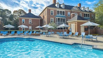 A swimming pool with lounge chairs and umbrellas, surrounded by a brick building with large windows and a covered porch.