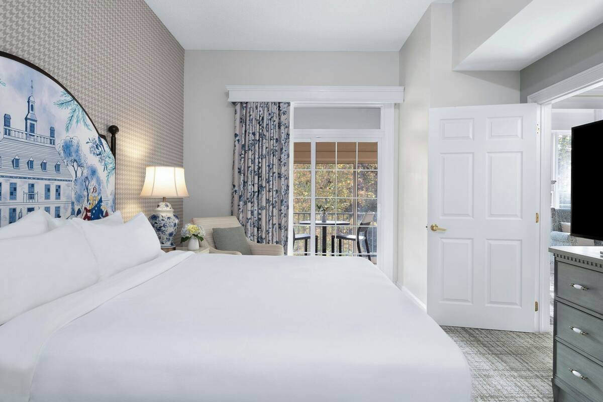A hotel room with a large bed, a desk with a lamp, a chair, a window with curtains, and a door.