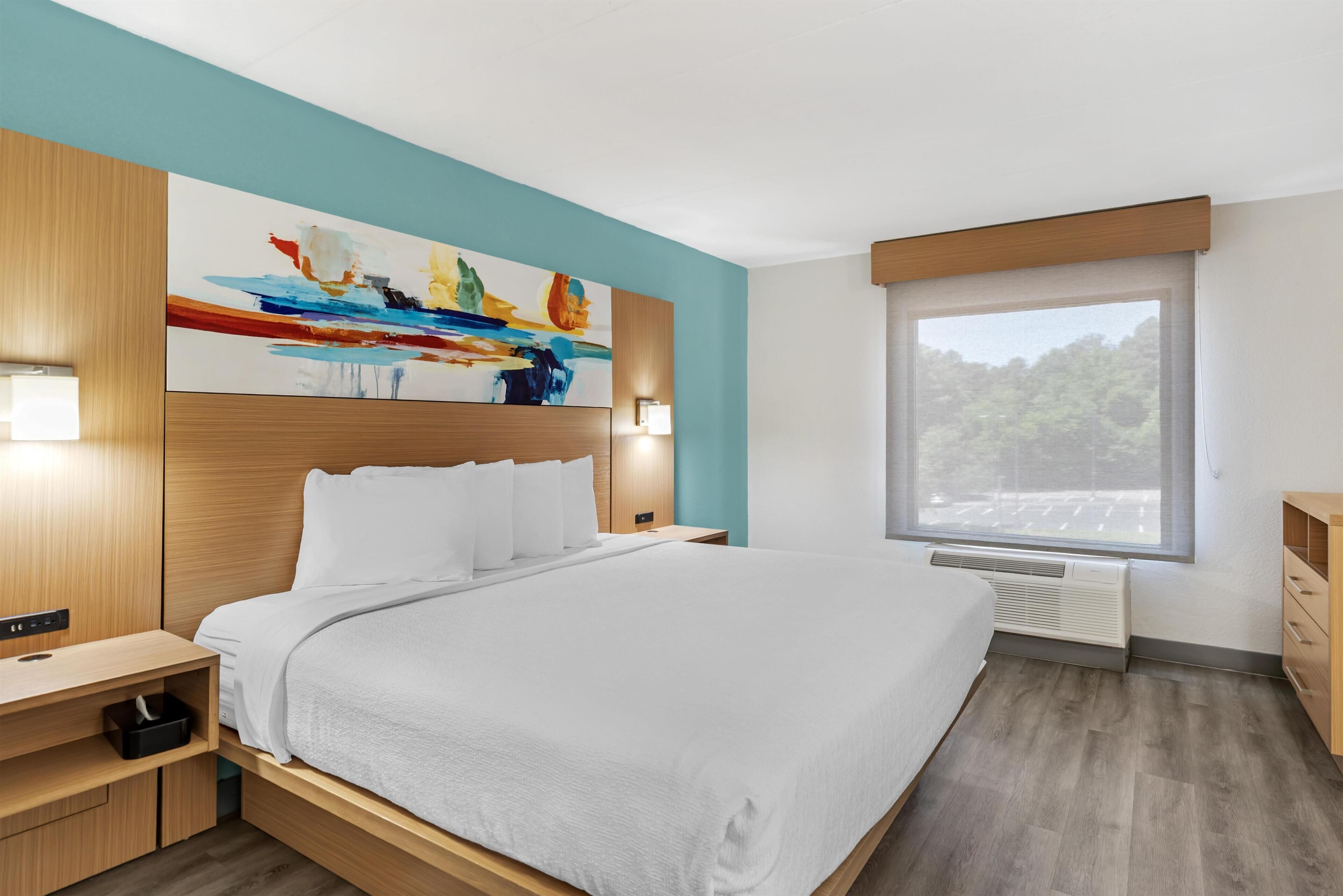 Una habitación de hotel moderna con una cama grande, vistas al exterior, un cabecero de madera y un cuadro en la pared.