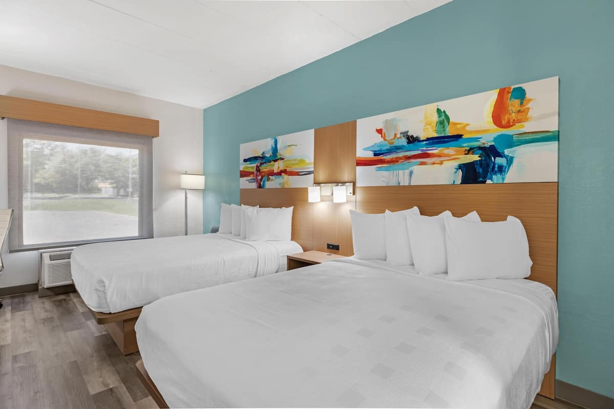 A hotel room with two beds, a large window, a painting on the wall, and a lamp on the nightstand.