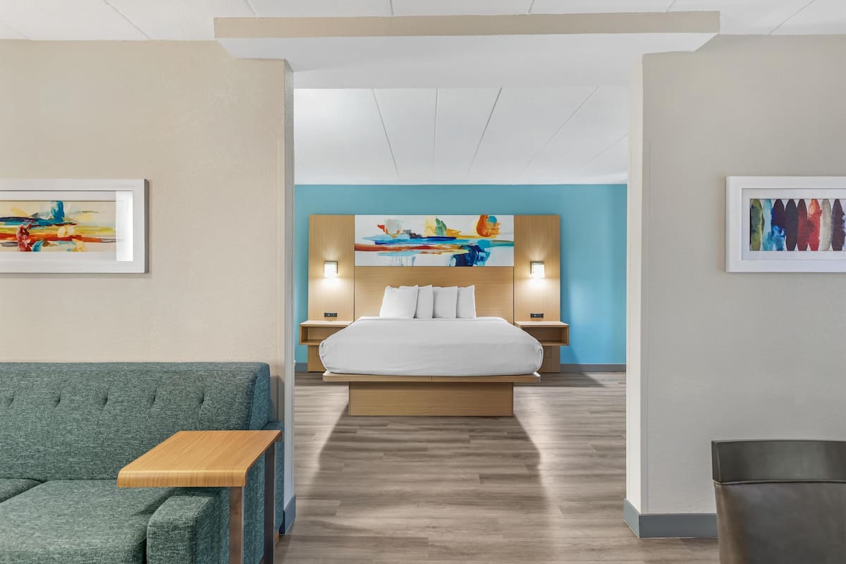 A modern hotel room with a large bed, a sofa, a wooden side table, and artwork on the walls.