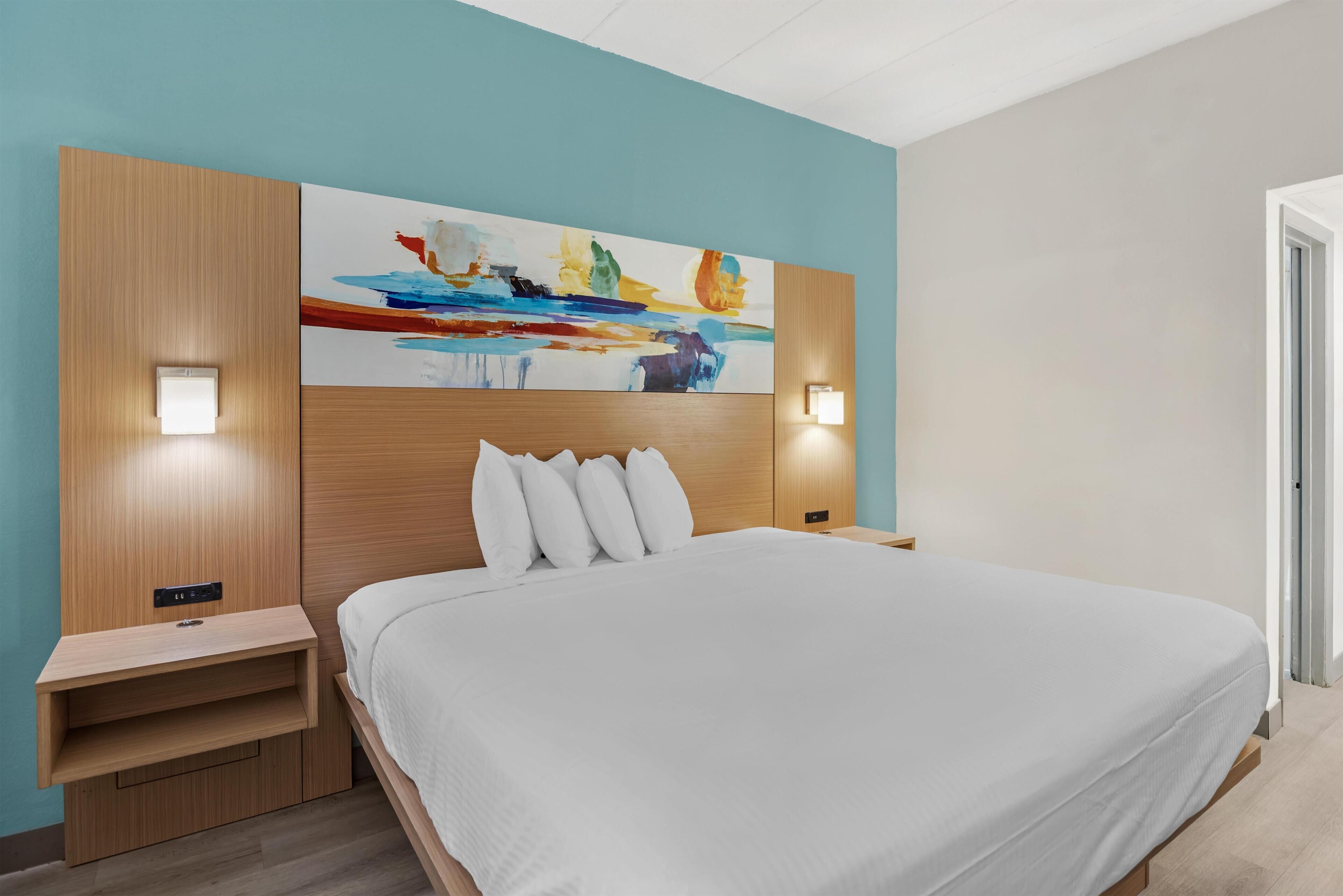 Una habitación de hotel moderna con una cama grande, cabecero de madera, dos mesitas de noche y un cuadro abstracto de colores en la pared.
