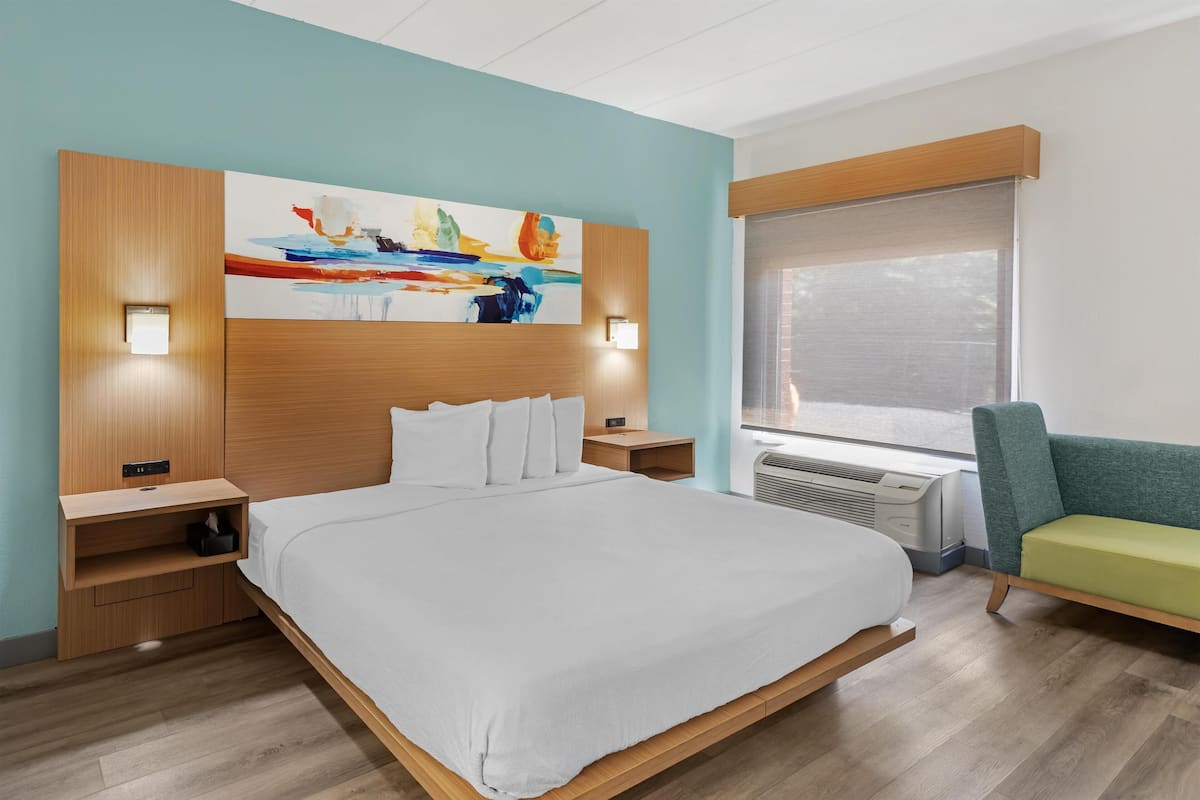 A modern hotel room with a large bed, bedside tables, a painting on the wall, a window with blinds, and a green chair.