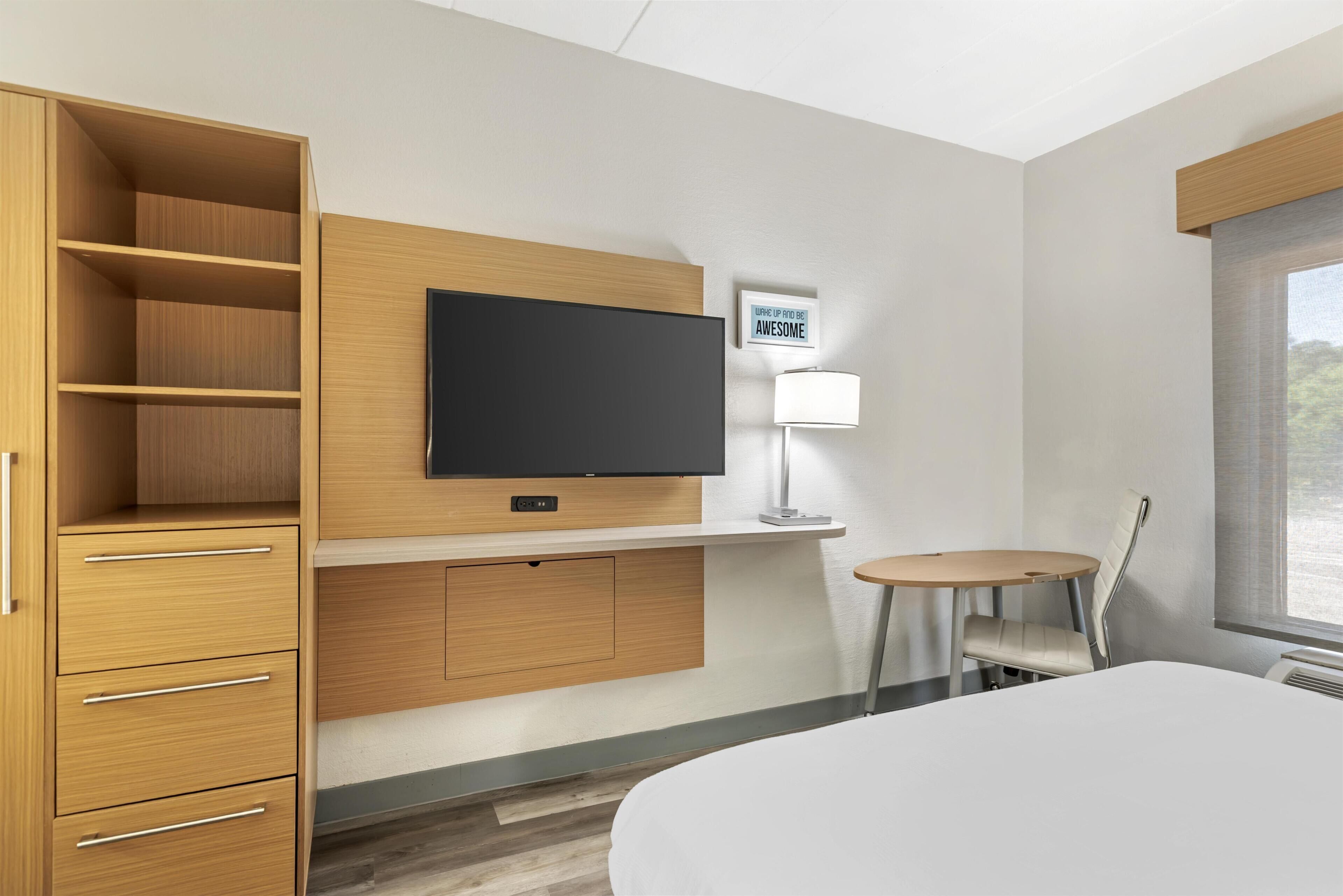 Quarto de hotel com uma cama, uma televisão fixada em um painel de madeira, uma mesa pequena com uma cadeira e uma janela com vista.