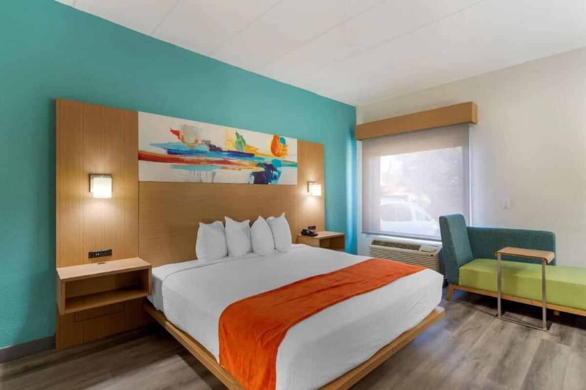 A hotel room with a bed, a nightstand, a chair, a window, and a painting on the wall.
