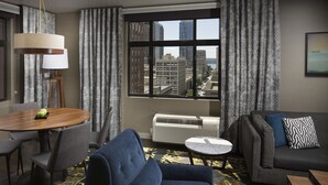 A hotel room with a dining area, a sofa, a small table, and a large window offering a city view.