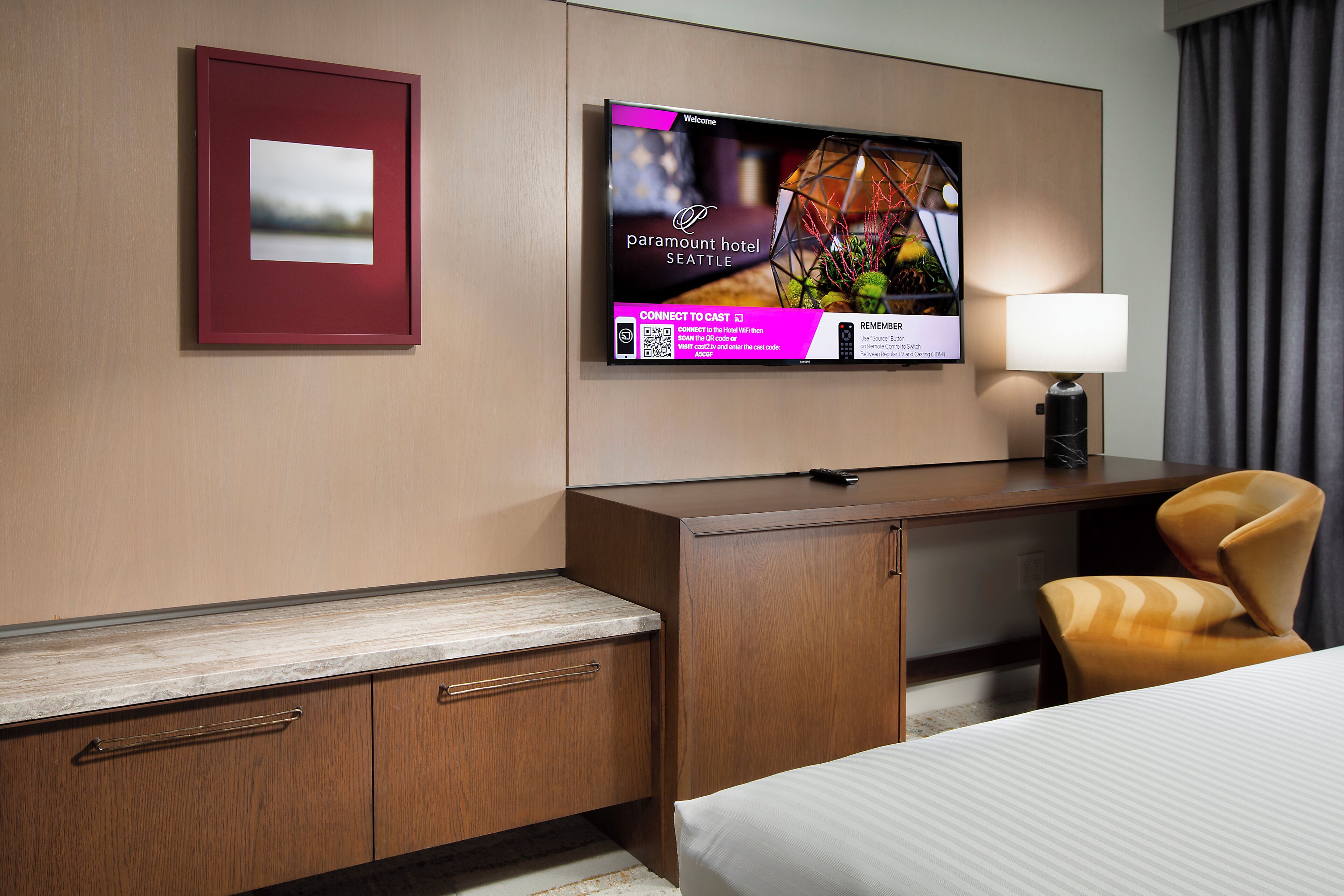 Ein Hotelzimmer mit einem Bett, einem Schreibtisch, einem Sessel, einem an der Wand befestigten Fernseher und einem gerahmten Bild.