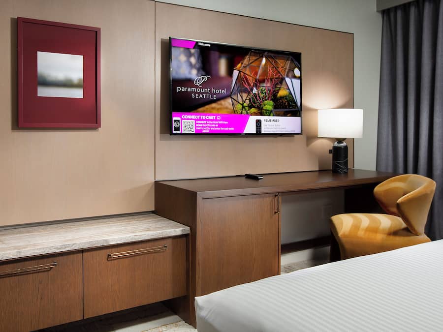 Habitación de hotel con cama, escritorio, silla, televisor montado en la pared y un cuadro enmarcado.
