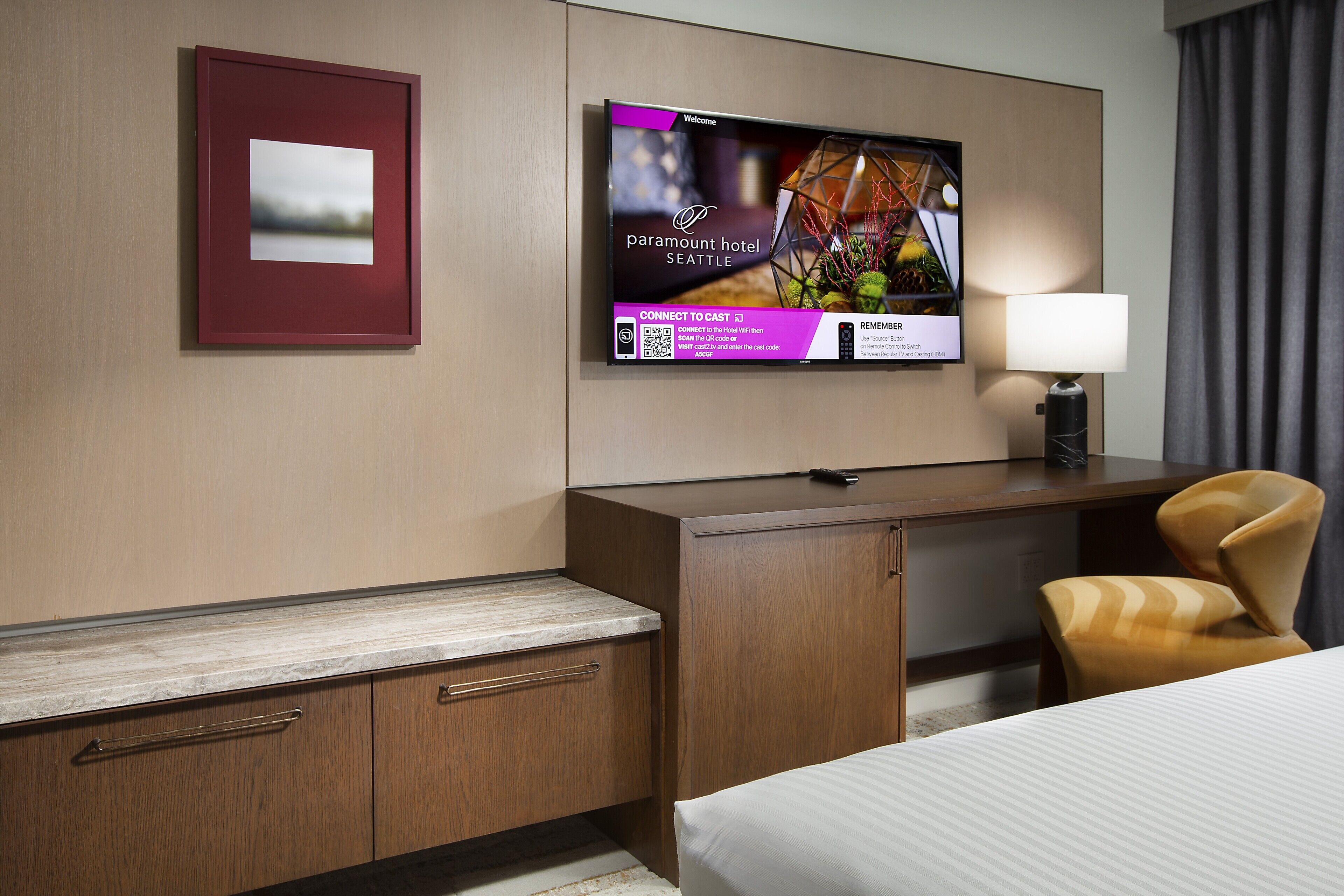 Habitación de hotel con cama, escritorio con lámpara, televisor mostrando un mensaje de bienvenida y un cuadro en la pared.
