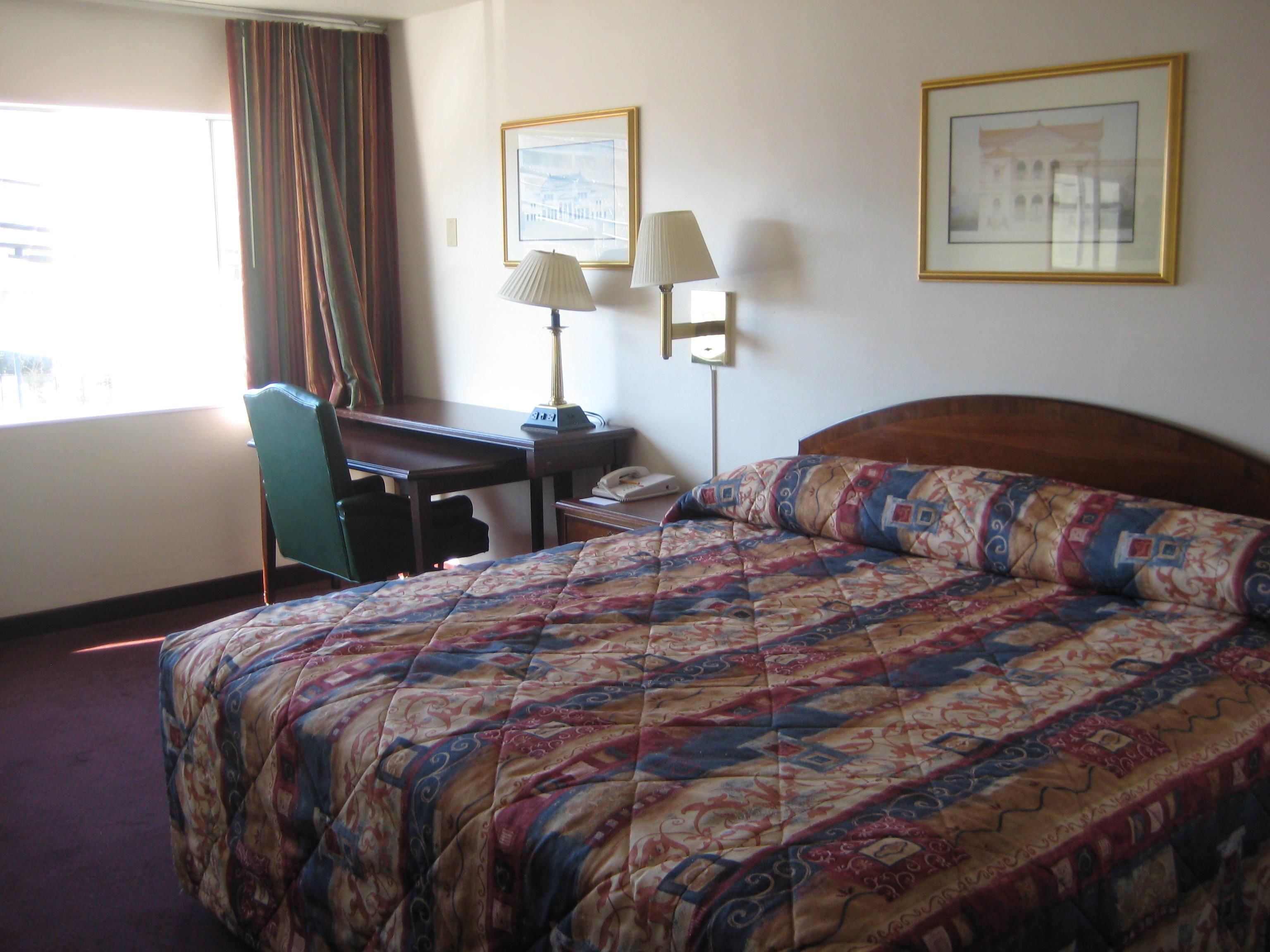 A hotel room with a bed, a desk, a chair, two lamps, and two framed pictures on the wall.