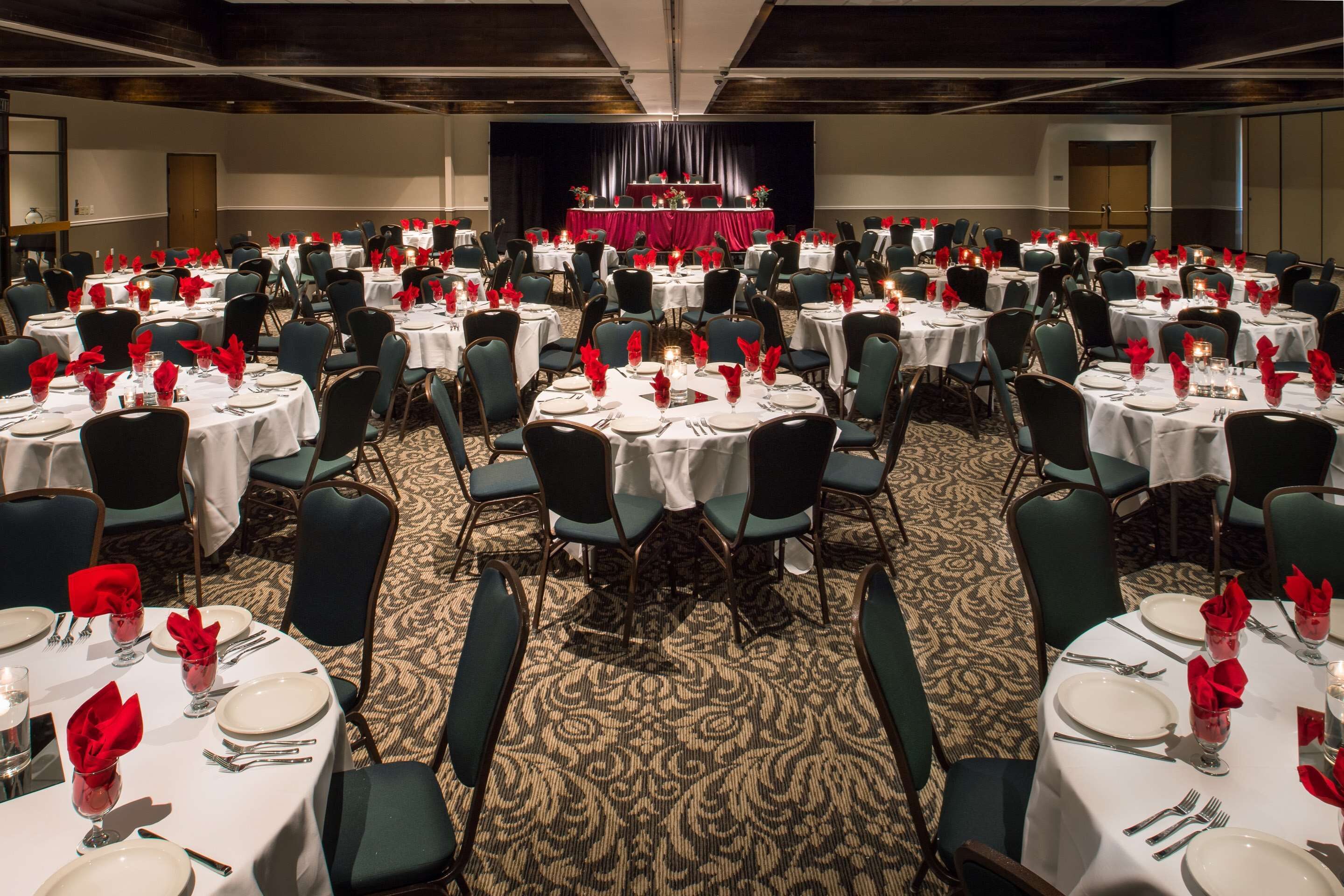 Un salón de banquetes con mesas redondas dispuestas para comer, cada una con mantel blanco, servilletas rojas y centros de mesa con velas.