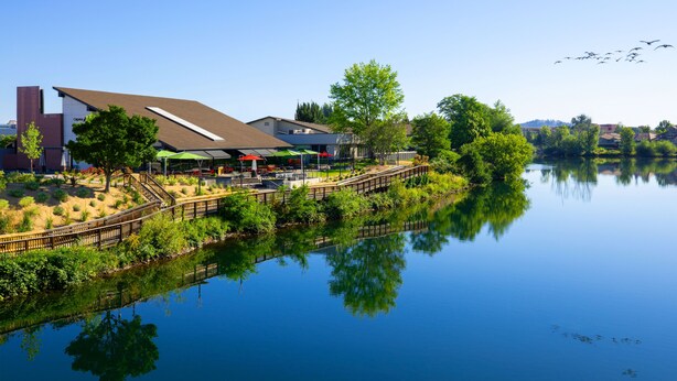 A lakeside view of a building with a covered outdoor area, a wooden bridge, and a reflection in the water.