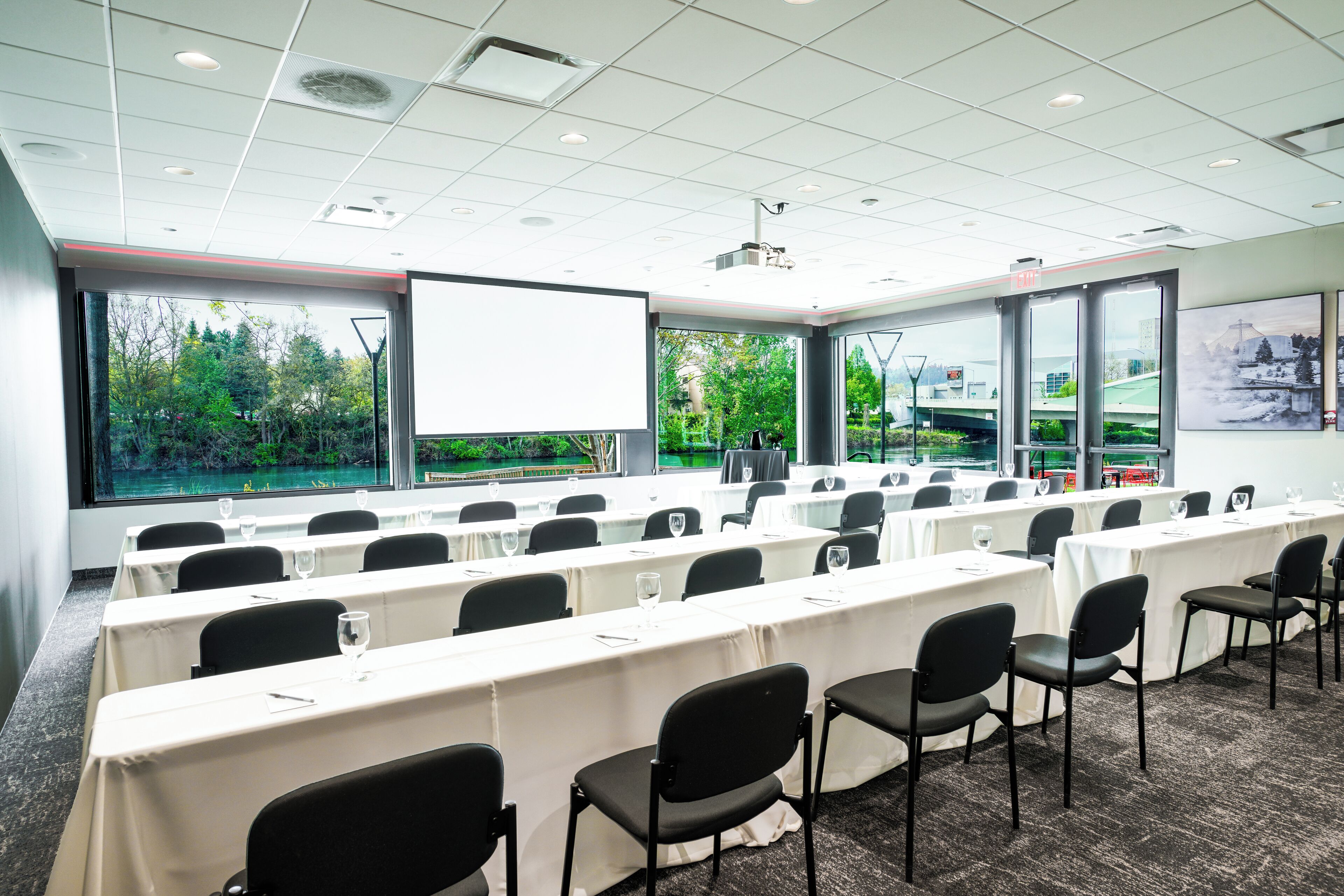 A conference room with tables and chairs, large windows with a view of trees, and a projector screen.