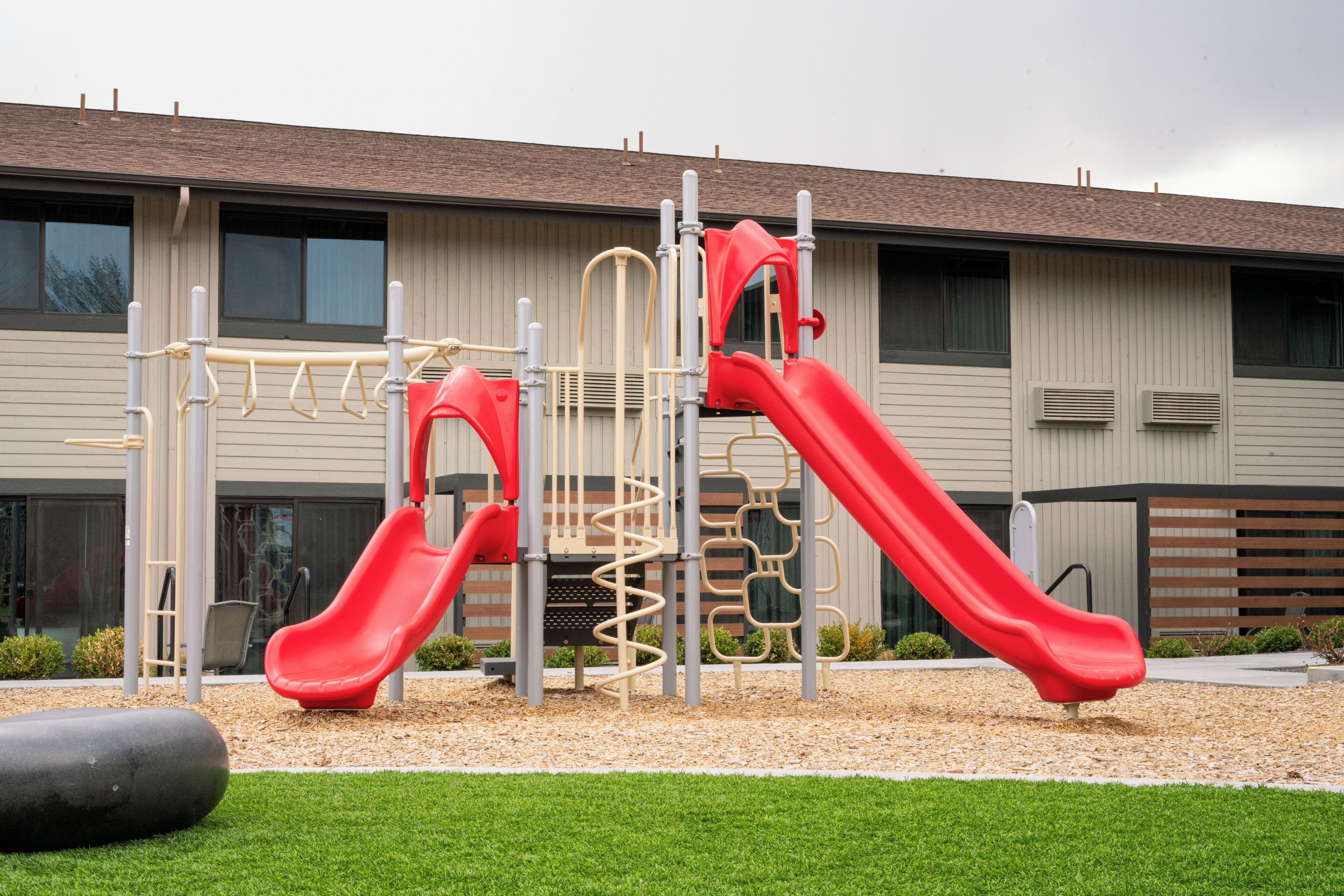 A playground with two red slides and a climbing structure, set against a building with large windows and air conditioning units.