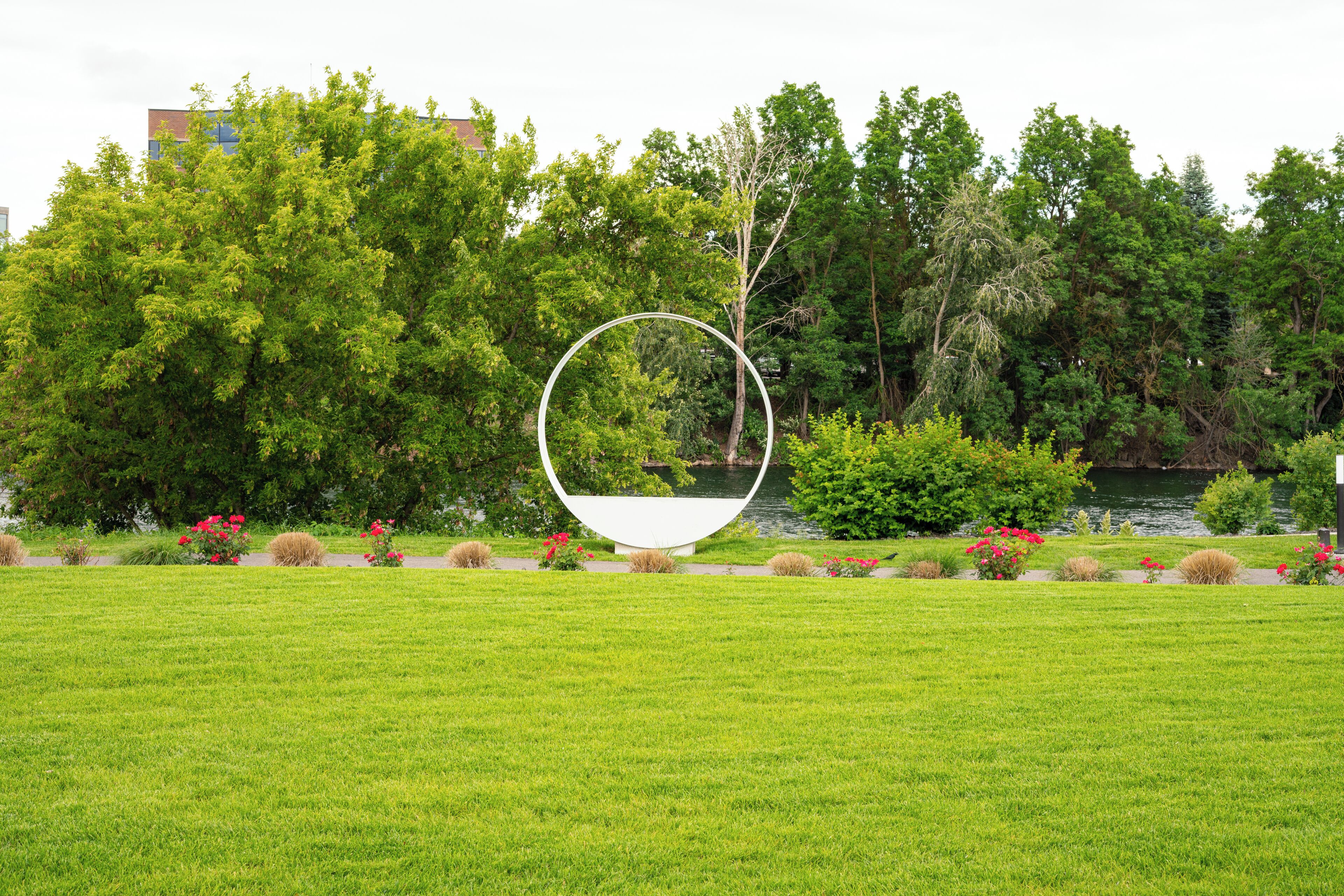 A park with a large circular sculpture in the center, surrounded by lush green grass and trees.