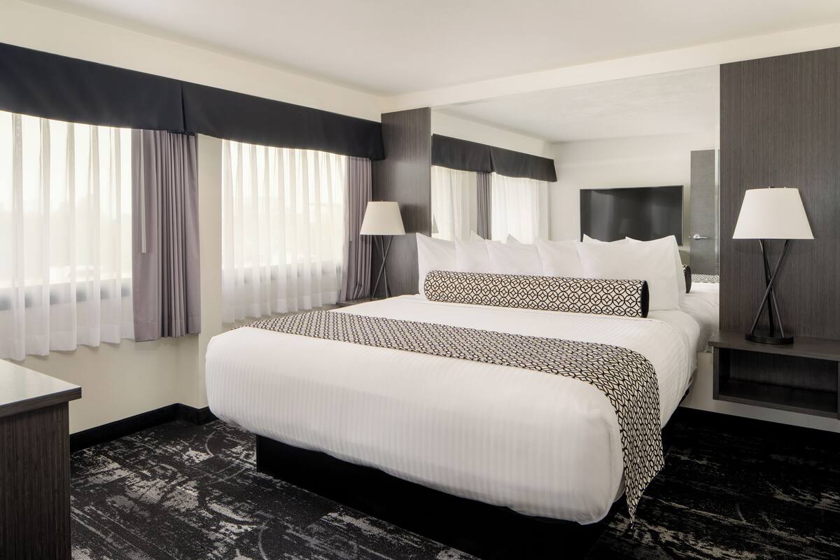 A modern hotel room with a large bed, two bedside lamps, a nightstand, and a large window with curtains.