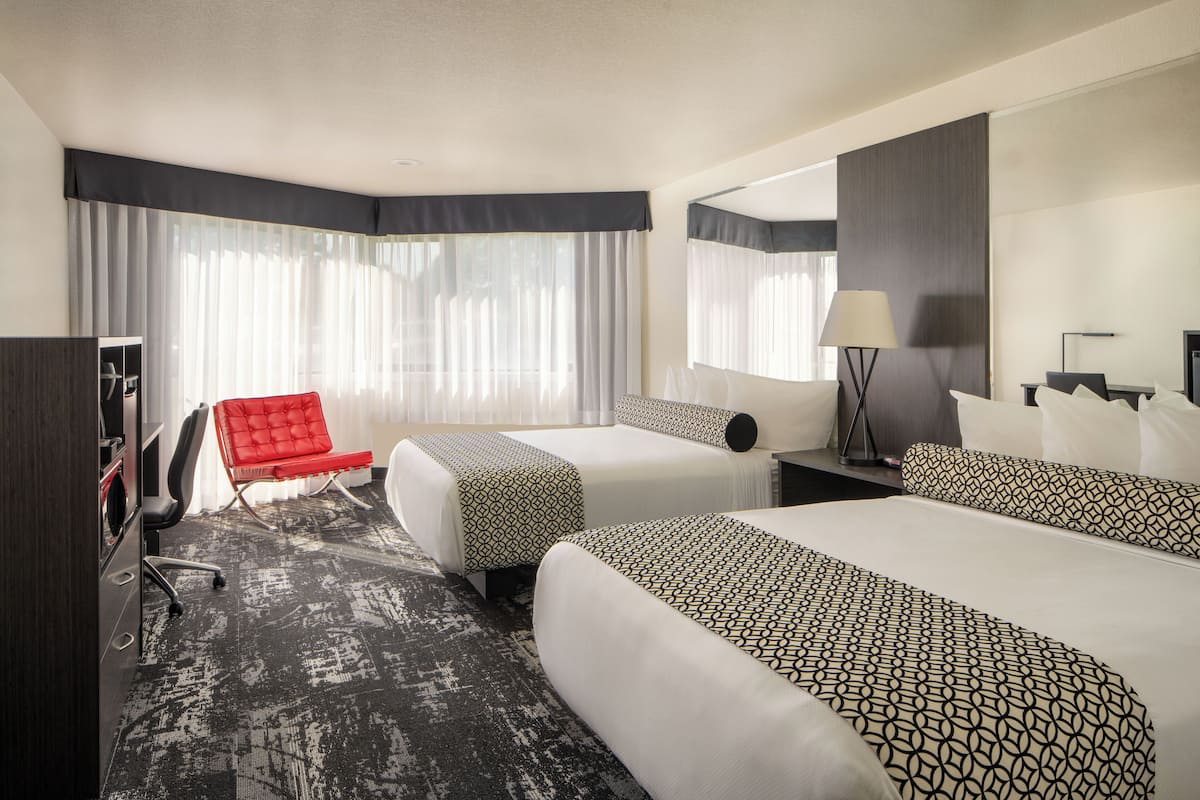 A hotel room with two beds, a red chair, a desk with a lamp, and a large mirror.