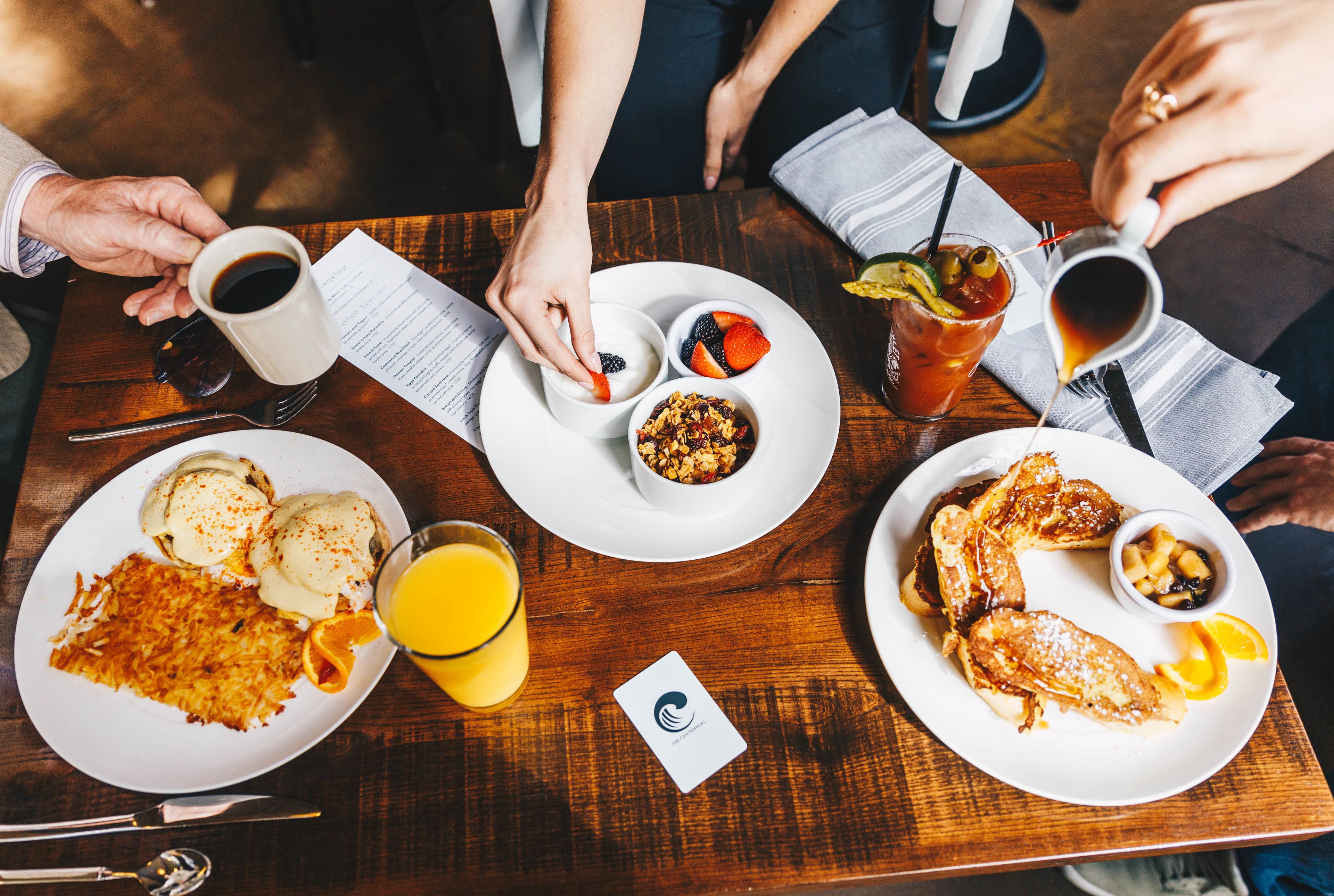 A wooden table with breakfast plates containing pancakes, eggs, and orange juice.