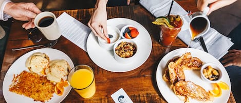 A wooden table with breakfast plates containing pancakes, eggs, and orange juice.