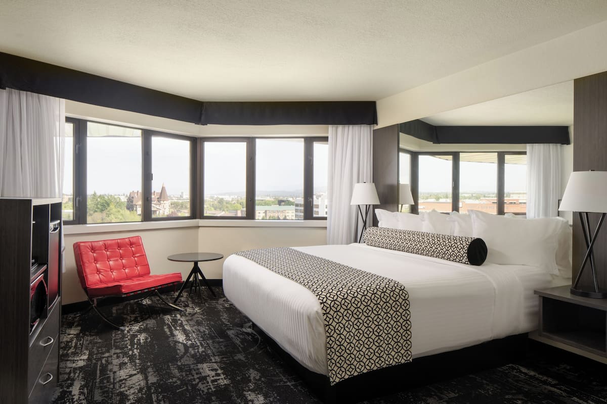 A hotel room with a large bed, a red chair, a small table, and a view of the city through large windows.