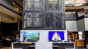 En moderne hotellobby med en receptionsdisk, to store skærme der viser bybilleder, og en dekorativ metalvæg.