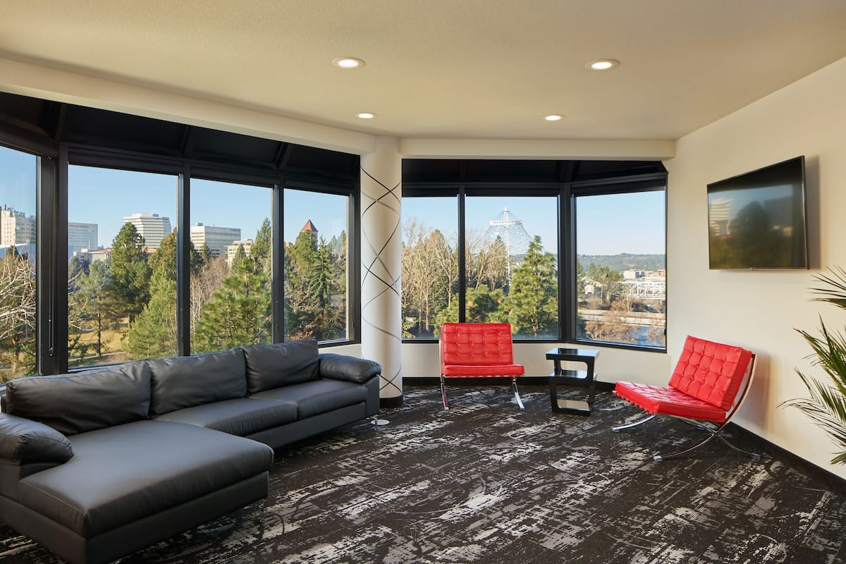 A modern room with a gray sectional sofa, a red chair, and a small table. Large windows offer a view of a cityscape and trees.