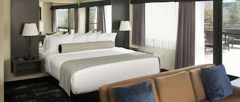 Habitación de hotel con una cama grande, dos mesitas de noche con lámparas, un sofá y vistas al exterior.
