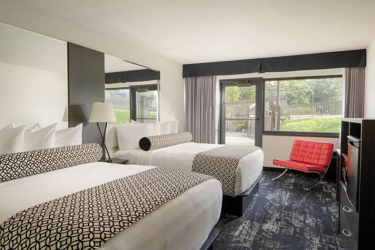 A hotel room with two beds, a red chair, a TV, and a large window with a view of the outdoors.