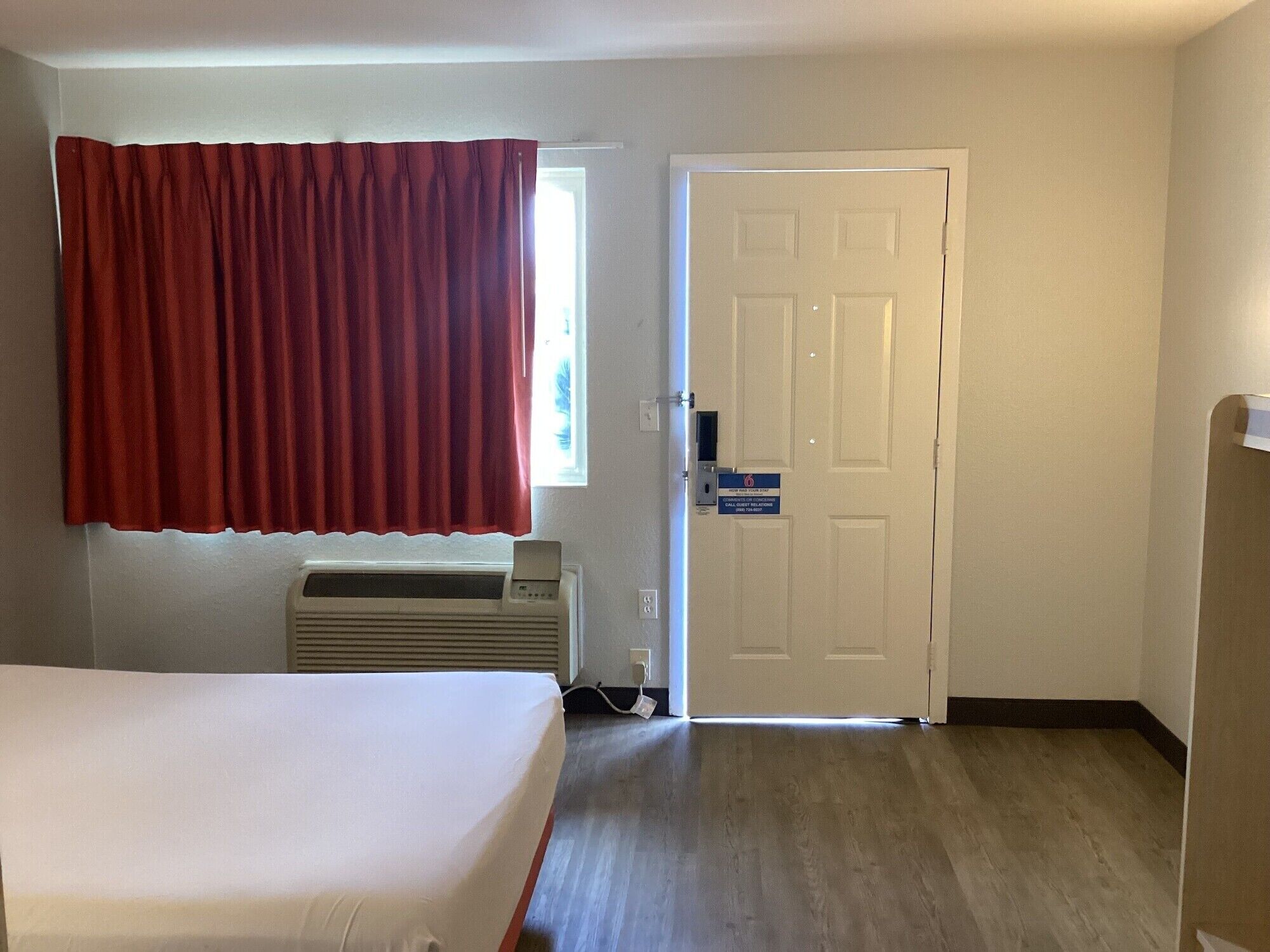 A room with a bed, a heater, a door with a sign, and a window with red curtains.