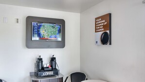 A room with a television mounted on the wall displaying a weather map, a coffee machine, and a sign advertising a morning charge-up for no charge.