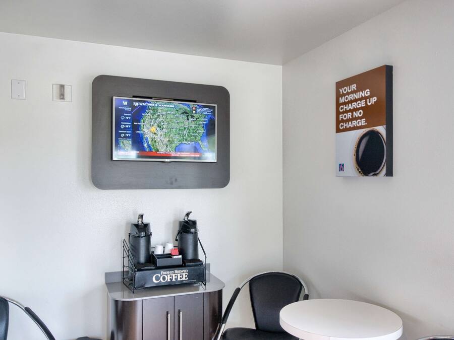 A room with a television mounted on the wall displaying a weather map, a coffee machine, and a sign advertising a morning charge-up for no charge.