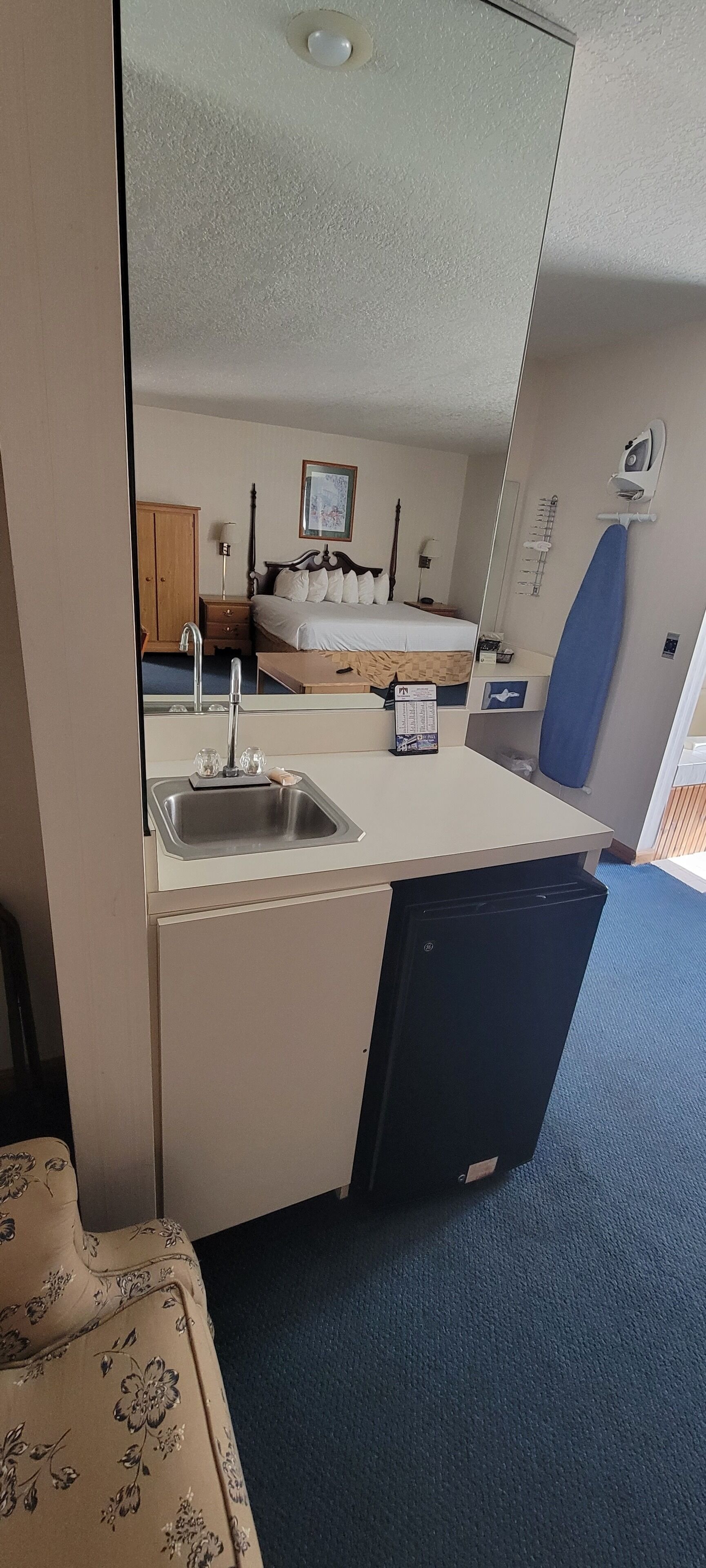 A compact room with a kitchenette, a bed, a mirror, and a small ironing board.