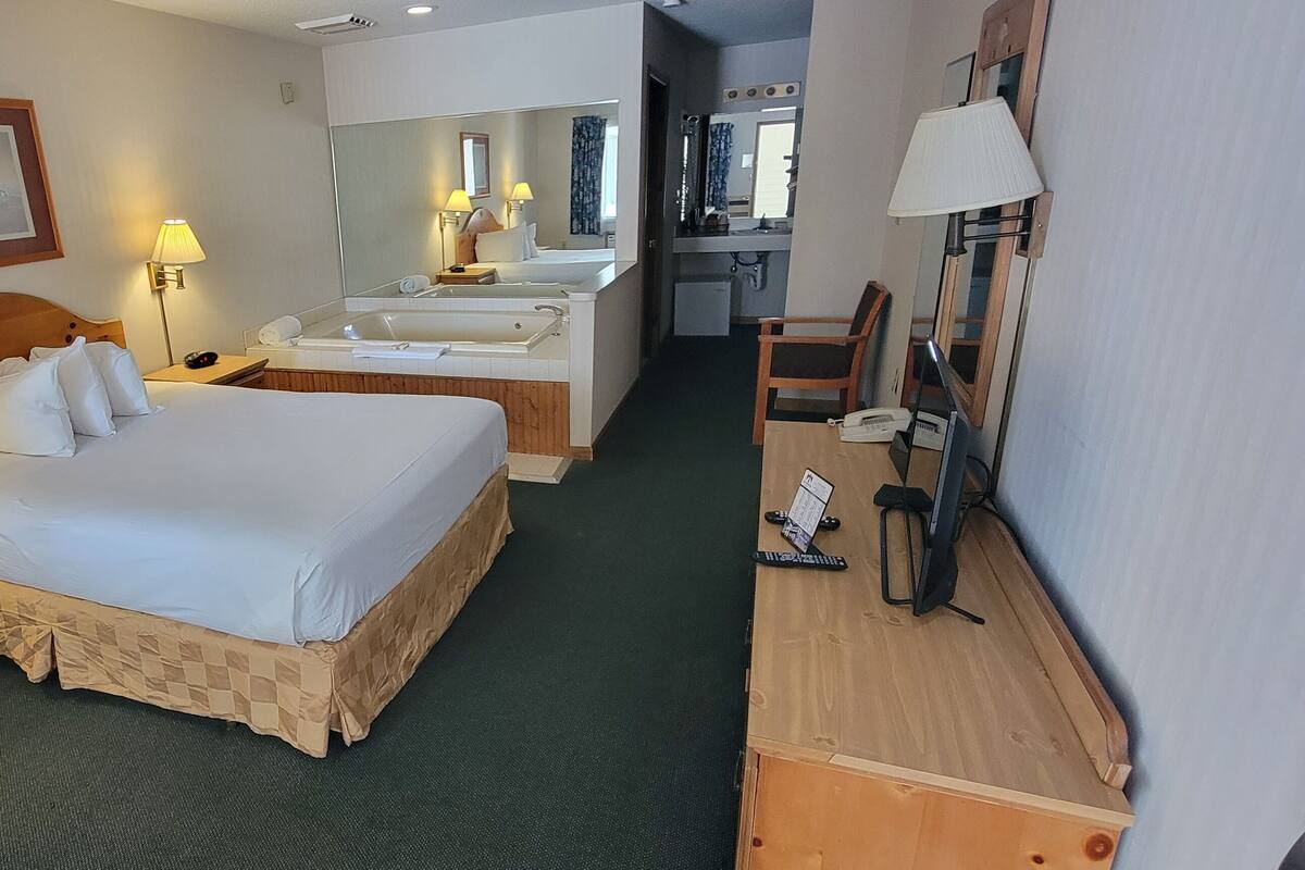 A hotel room with a large bed, bedside tables, a desk with a chair, a mirror, and a bathroom with a bathtub and sink.