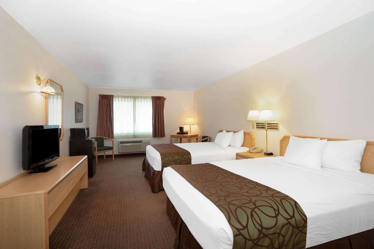A hotel room with two beds, a desk with a TV, a chair, a desk lamp, and a bedside table with a lamp.