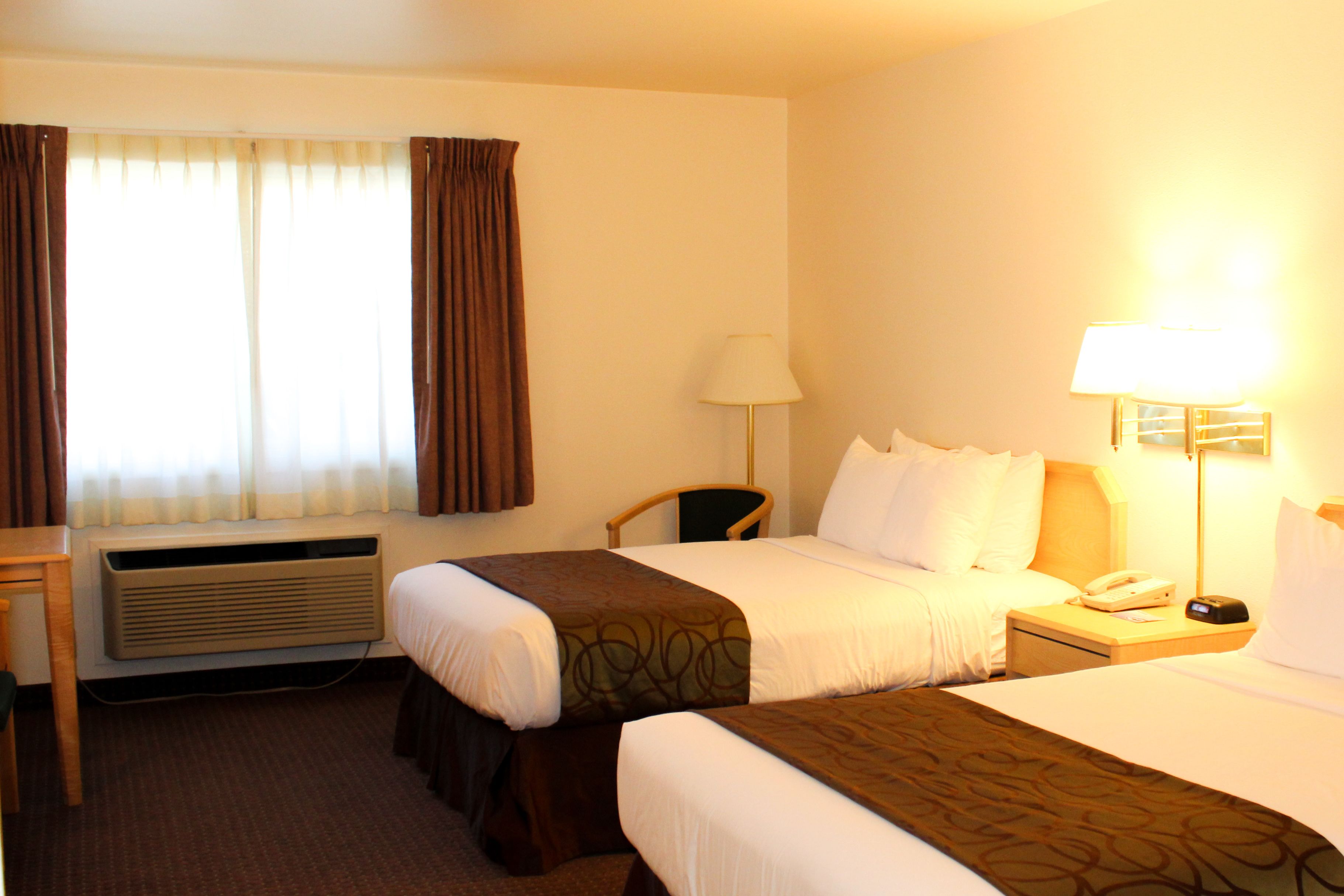 A hotel room with two beds, a desk, a chair, a lamp, a phone, and a window with curtains.