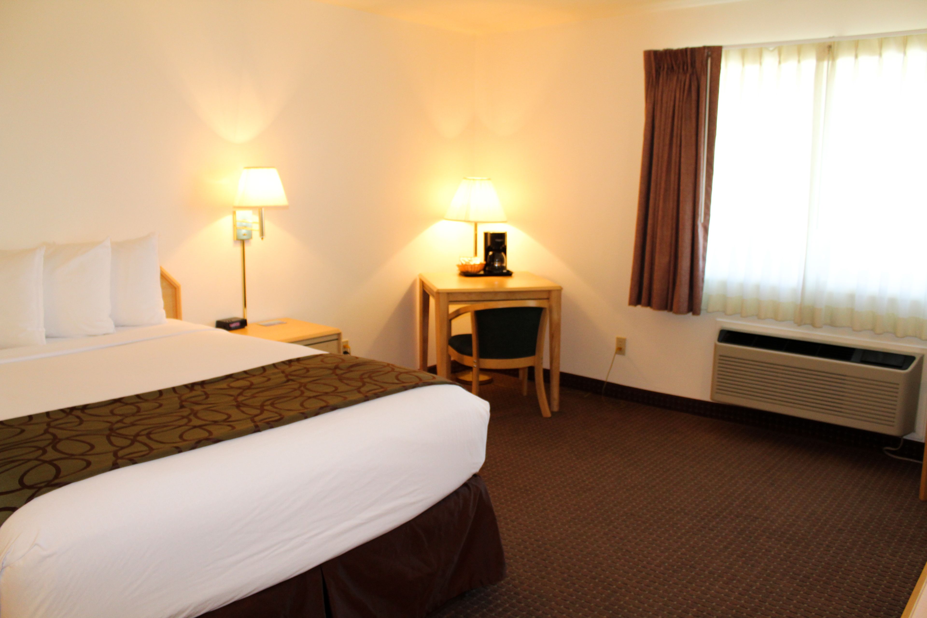 A hotel room with a large bed, a desk with a lamp, a chair, and a window with curtains.