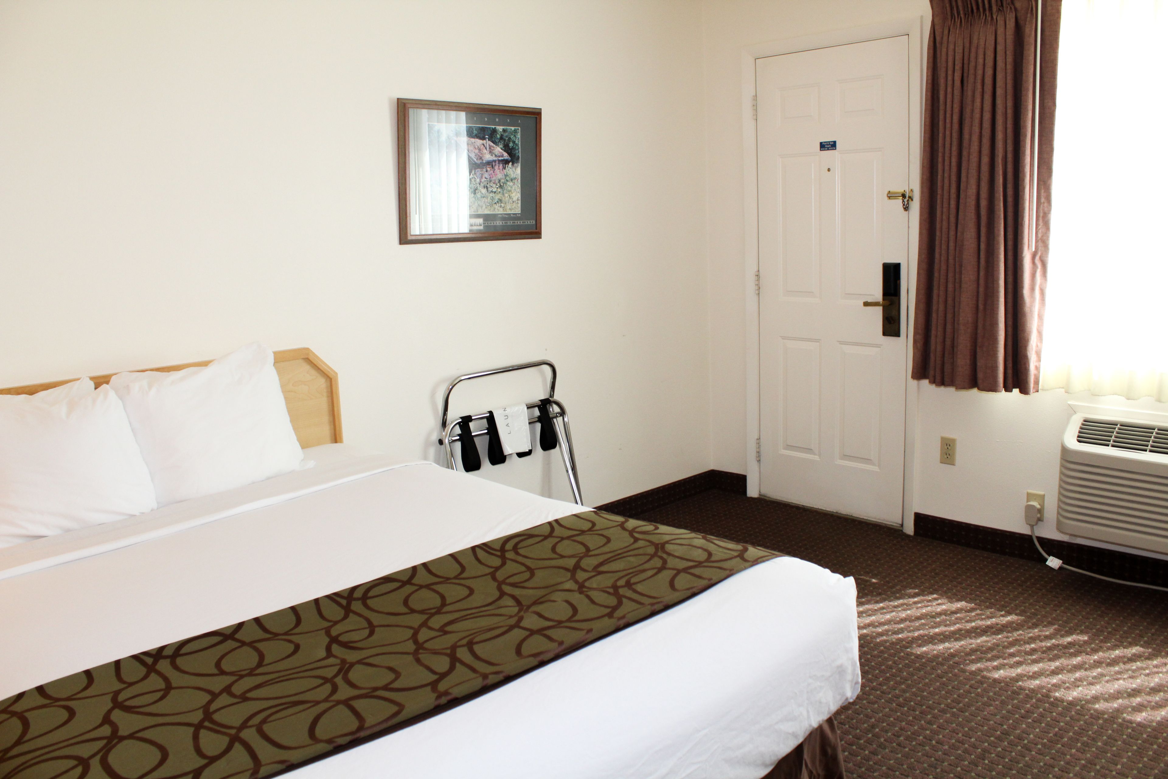 A hotel room with a bed, a picture on the wall, a door, and an air conditioning unit.
