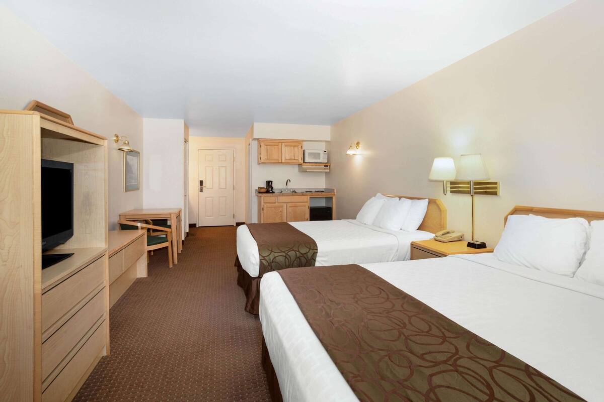 A hotel room with two beds, a desk, a TV, a kitchenette, and a bathroom.