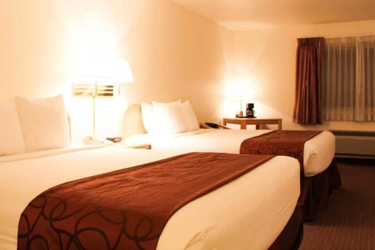 A hotel room with two beds, a bedside table with a lamp, a phone, and a window with curtains.