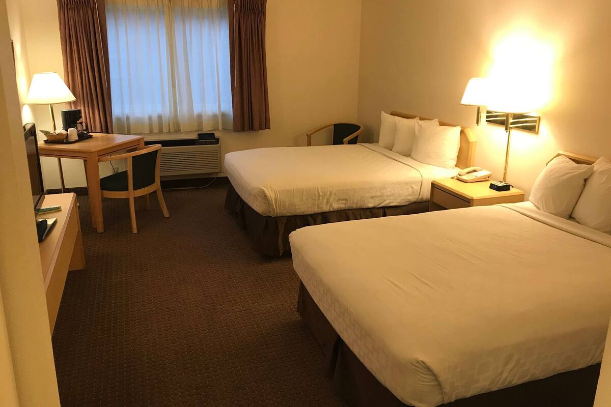 A hotel room with two beds, a desk, a chair, and a nightstand with a lamp.