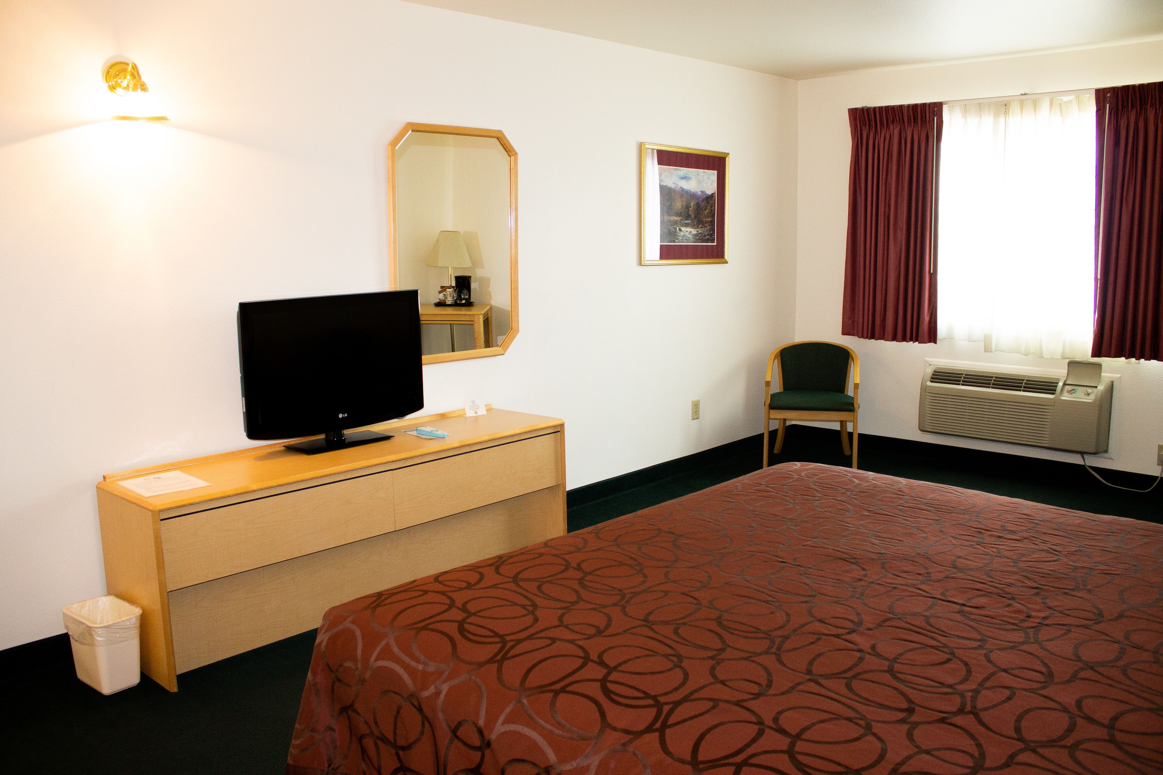 A hotel room with a bed, a TV on a wooden dresser, a chair, a mirror, a picture on the wall, and an air conditioning unit.